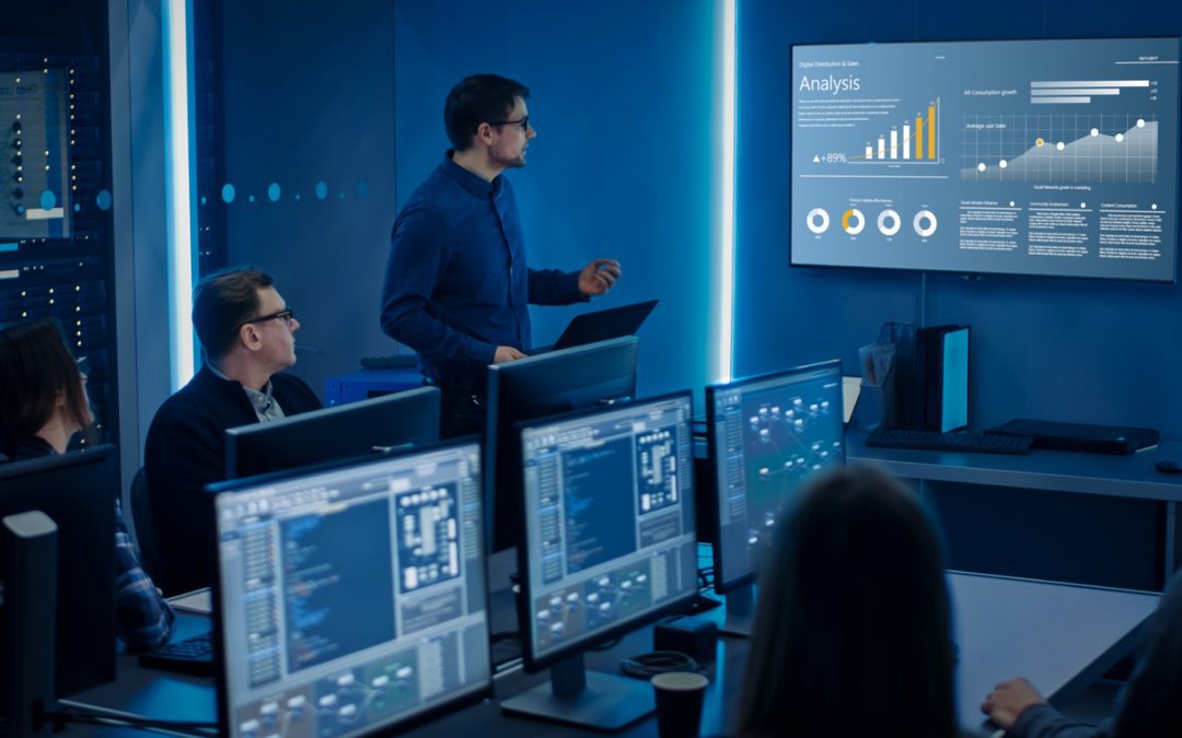 DevOps team at shared table in tech-centered room while analytics are cast onto wall-mounted screen